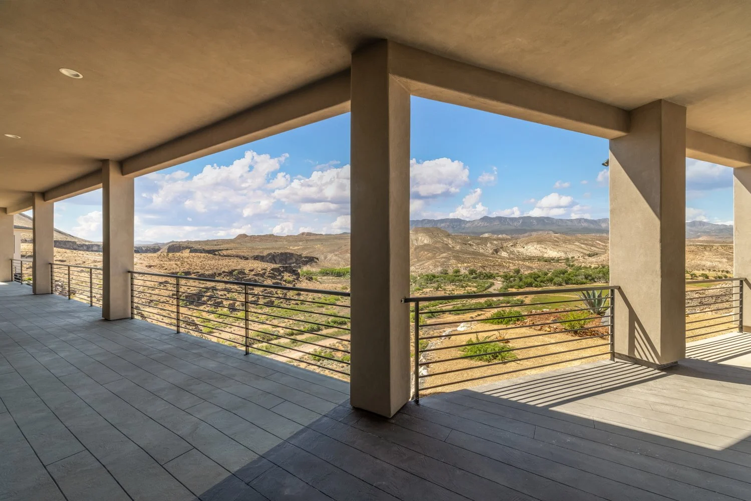 View from a covered balcony or terrace showing a desert landscape with mountains, scattered vegetation, and a cloudy sky.
