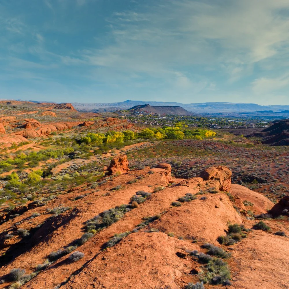 A panoramic view of a desert landscape with reddish rocks, sparse green vegetation, and a distant town or city under a partly cloudy sky.
