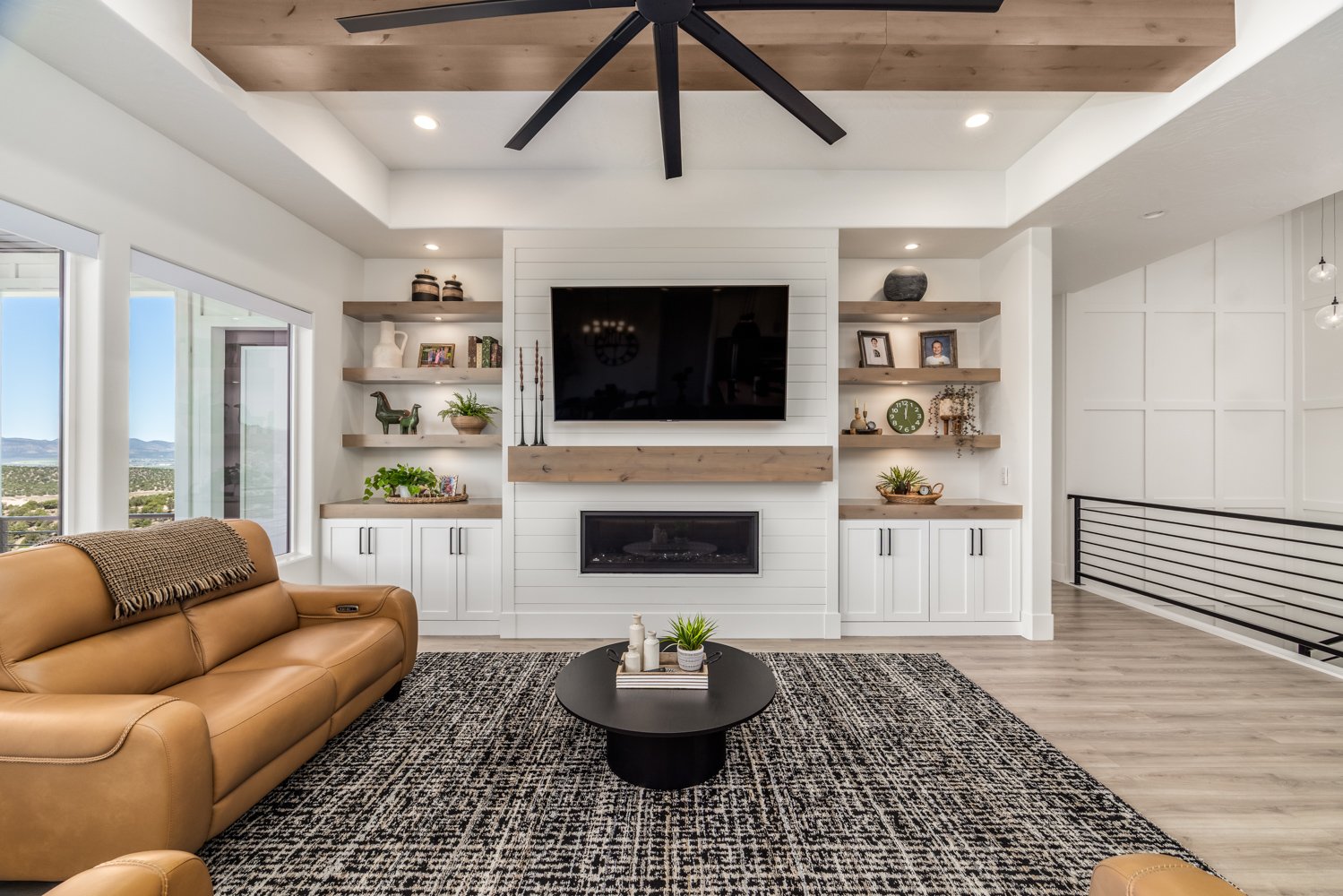 Living room with a large tan couch, black coffee table, and a wall-mounted TV above a fireplace, surrounded by built-in shelves with decorative items. Windows on the left side reveal a scenic view of the outdoors.