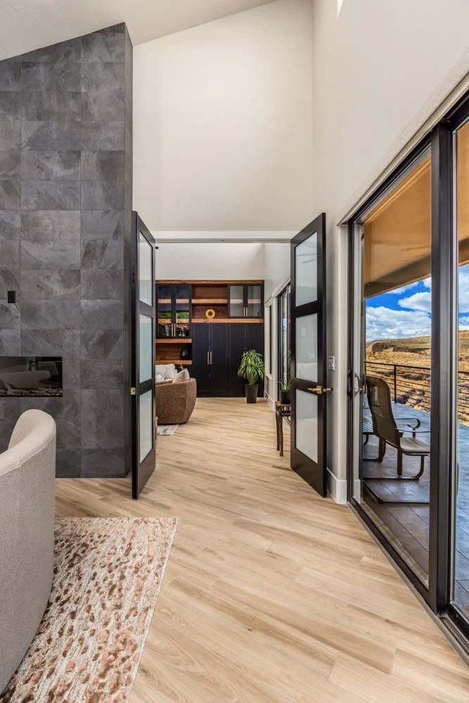 Interior of a modern house with a view of an outdoor patio and landscape through sliding glass doors, featuring wooden floors and contemporary furniture.