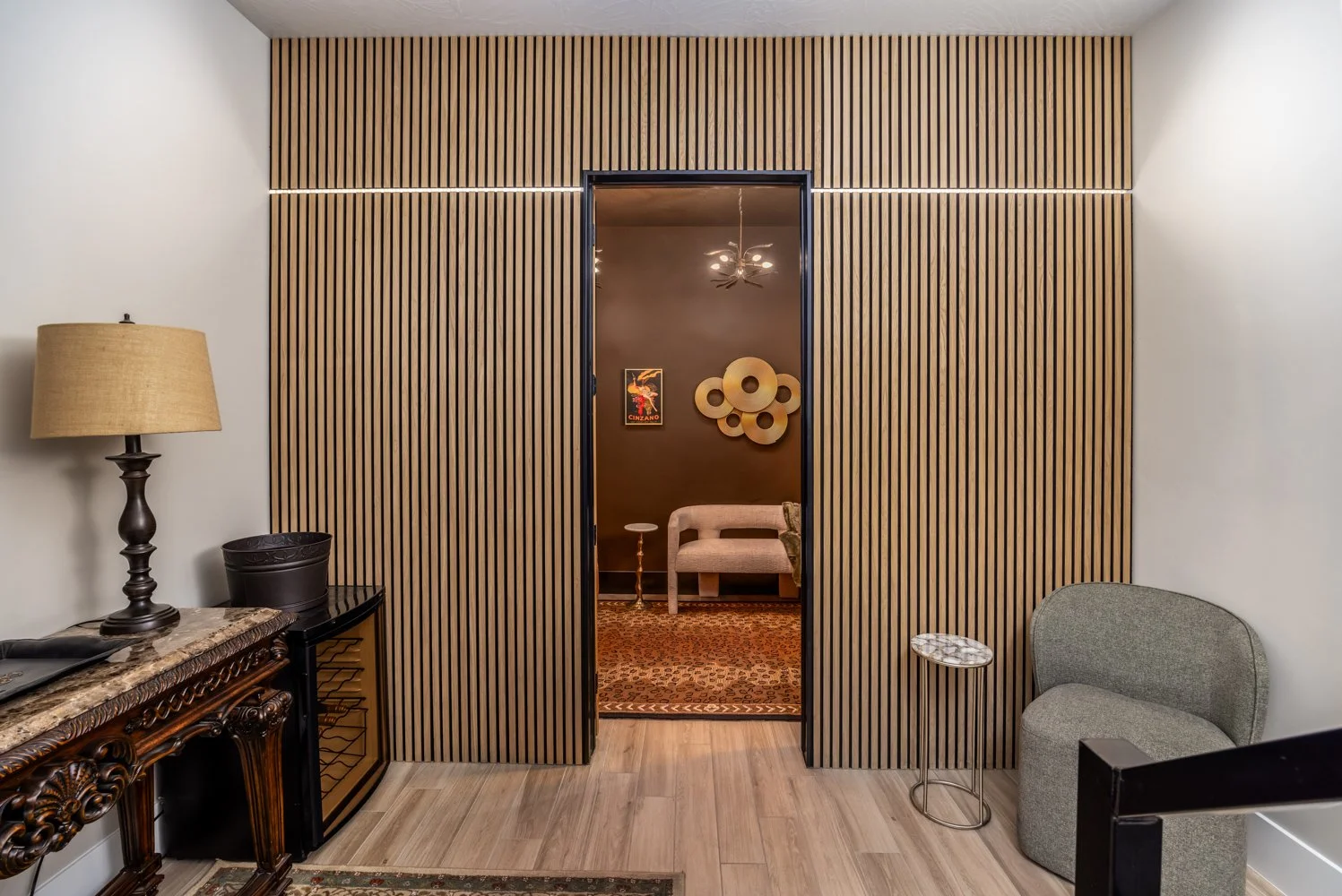 An interior view showing a wooden slat accent wall with a doorway leading to a room with dark walls and modern decor, including a beige chair, a small side table, and wall art. There is a vintage wooden console table with a lamp on it, a gray upholst