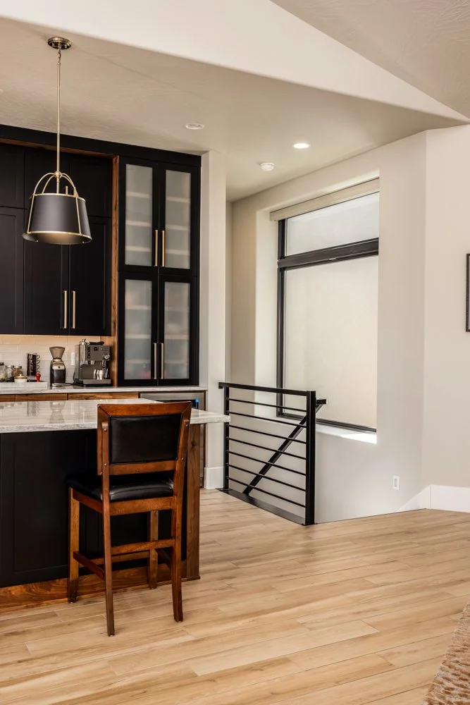 Modern kitchen with black cabinets, a marble countertop, a bar stool, large window, and a staircase railing.