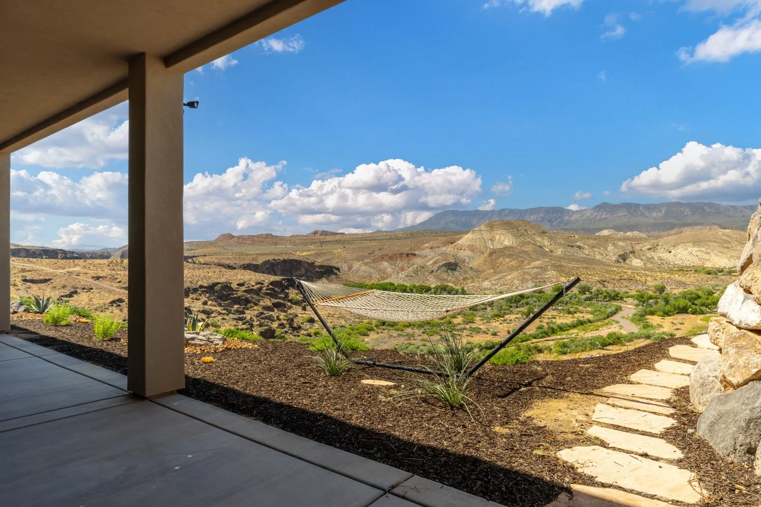 View from a patio featuring a hammock, stone pathway, desert landscape with sparse vegetation, mountains in the distance, and a partly cloudy blue sky.