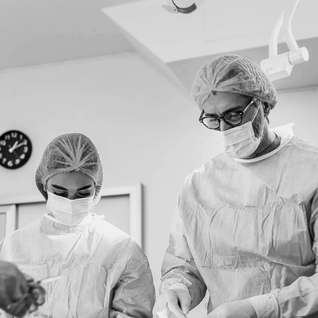 Two medical professionals in surgical attire, including masks, hair covers, and gowns, performing a procedure in an operating room.