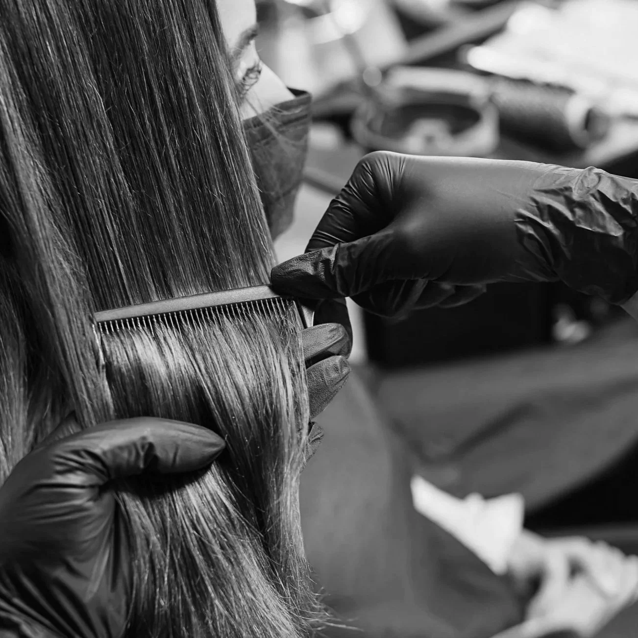 A hairdresser wearing black gloves uses a fine-tooth comb to precision work on a client's long, straight hair. The setting appears to be a salon with various tools and products visible in the background.