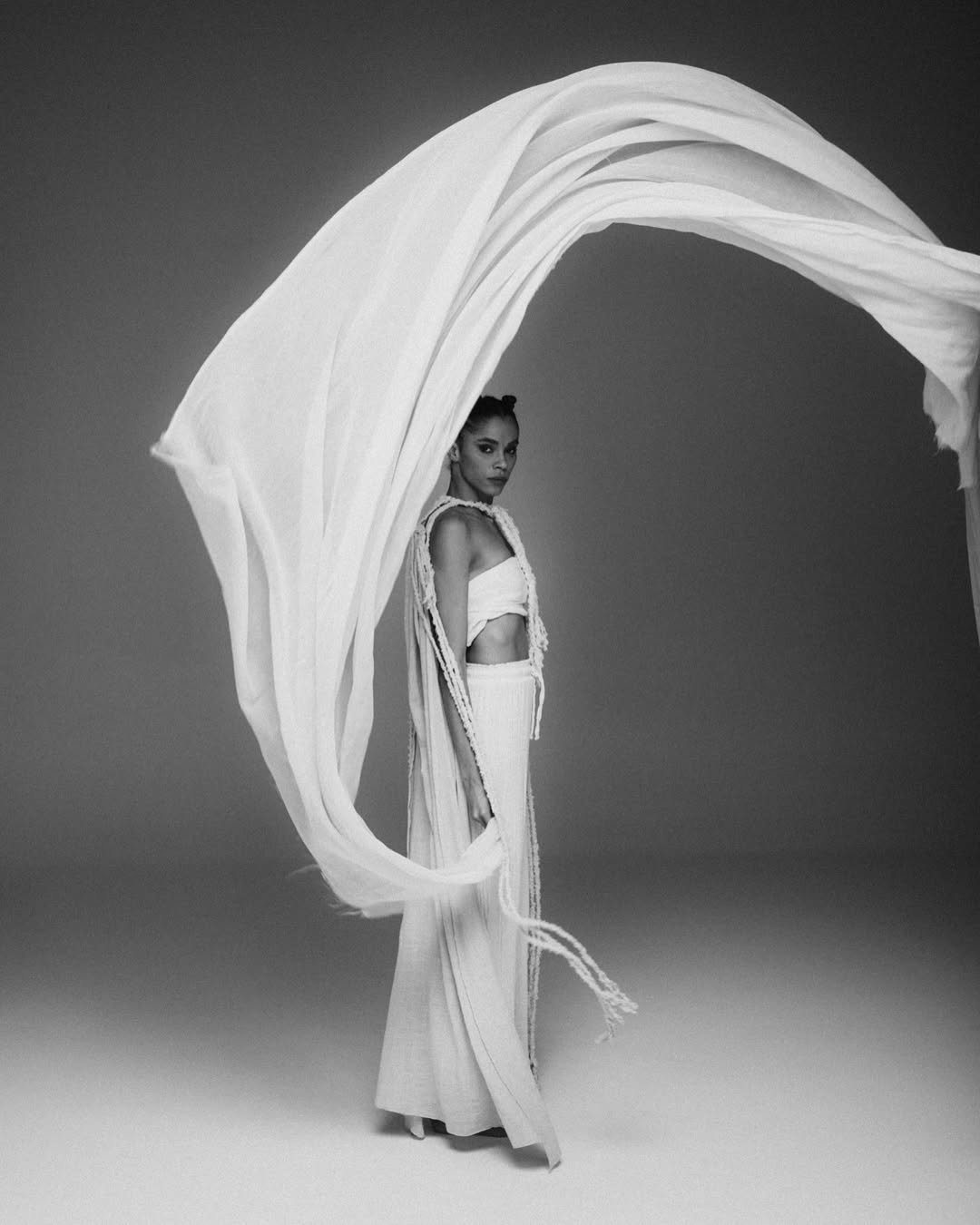 A woman in a white outfit with fringe, standing next to a large, flowing white fabric that curves around her, in a black-and-white studio portrait.