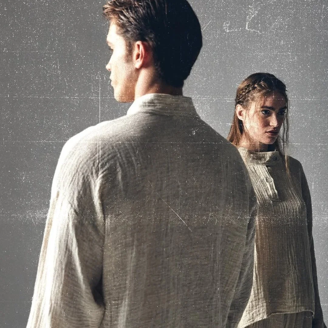 A man and a woman stand with their backs partially turned, facing away from each other, both wearing beige textured jackets against a plain background.