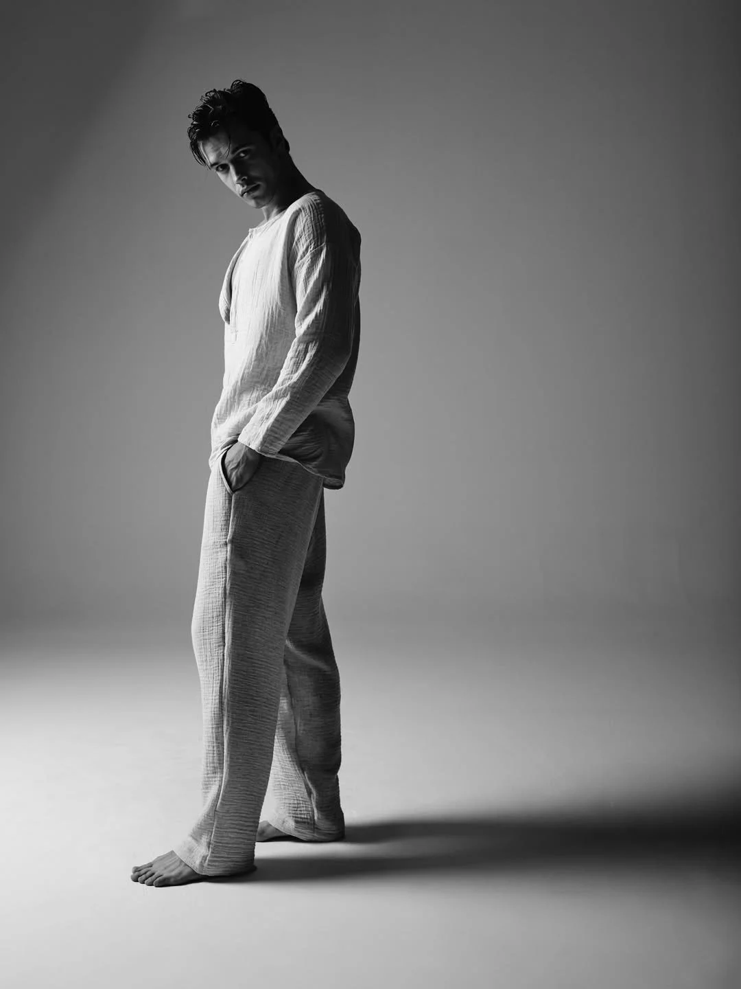 A young man standing on a plain background, dressed in light-colored loungewear with hands in pockets, gazing at the camera in a black-and-white photo.