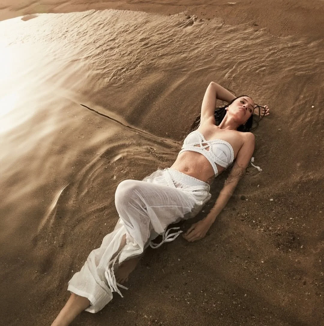 A woman wearing a white bikini top and white loose pants is lying on a sandy beach, partially covered with water, with her arm resting behind her head and eyes closed.