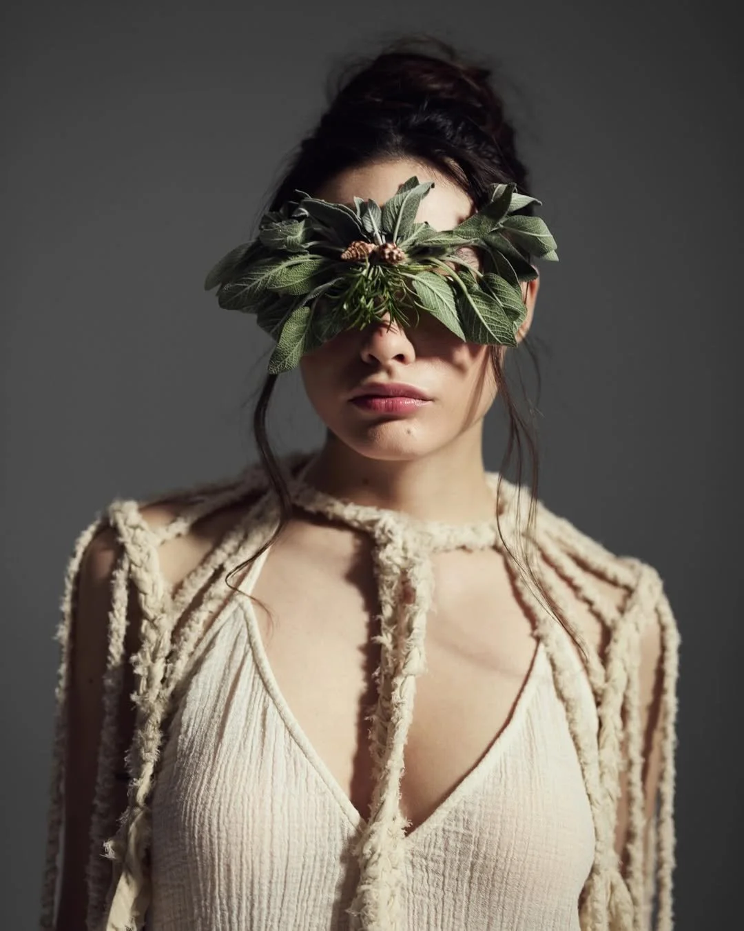 A woman with dark hair styled in loose waves, wearing a beige top and a textured, beige cardigan with cut-out shoulders. She has a leafy branch covering her eyes, creating a surreal and artistic appearance. The background is plain and gray.