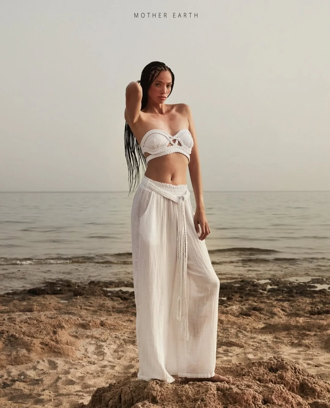 A woman in a white bikini top and flowy white pants standing barefoot on a sandy beach with the ocean in the background. The sky is overcast, and the woman has long dark braids.