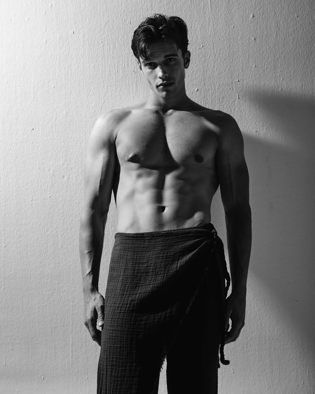 A shirtless young man standing against a plain wall, looking at the camera with a serious expression, with shadows casting on his left side.