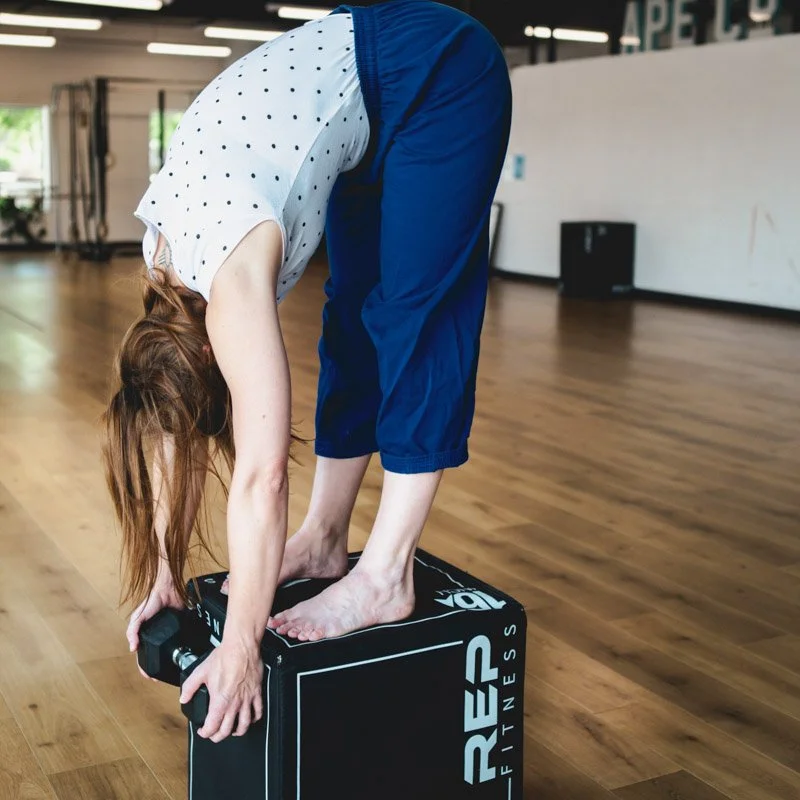 Leah is standing on a black fitness box, bent over with her whole spine and arms pulling towards the floow. She is holding a dumbbell with both hands and her hair is down. She is in a brightly lit movement studio.