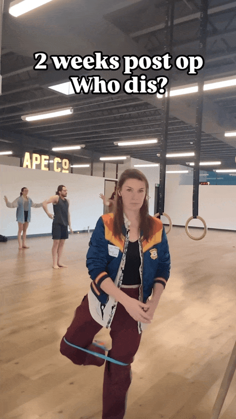 Leah in the foreground is doing a hip exercise with a resistance band. In the background, students take movement class. Text overlay reads "2 weeks post-op. Who dis?"