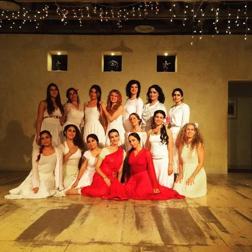 A group of women dancers in white and red attire pose for a photo in a studio with string lights overhead.