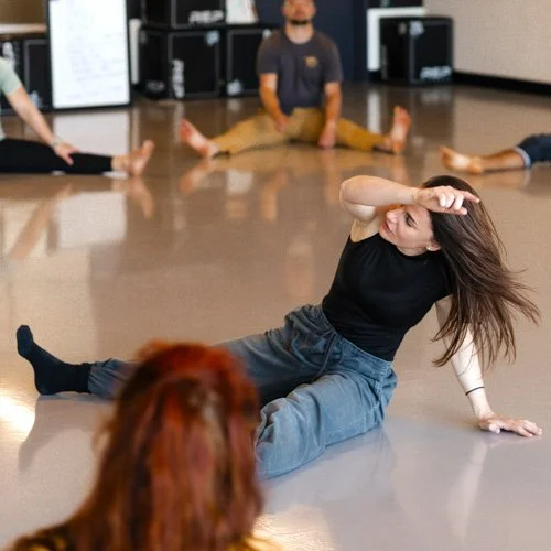 Leah a movement teacher with long brown hair is sitting on the floor, raising her arm above her head while she demonstrates a floorwork movement to a class with other participants in the background.