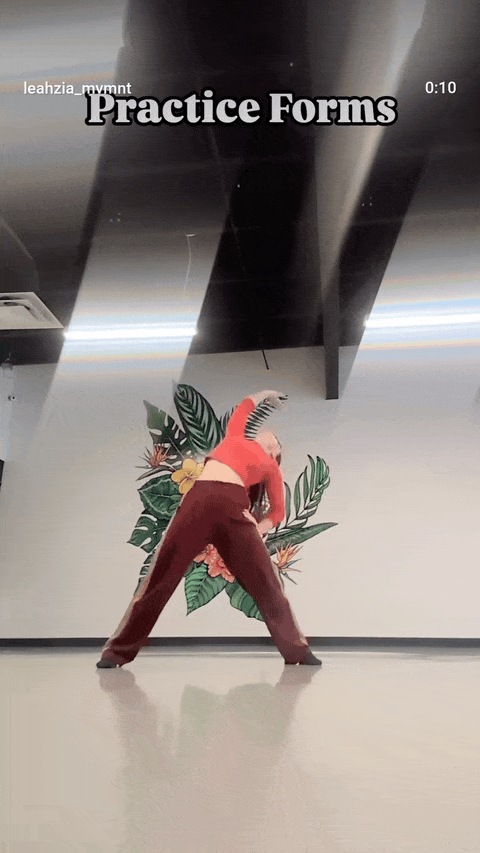 Leah in a red top and burgundy pants does stretching exercises in a Denver studio, with a mural behind them.