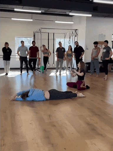 A group of people stand in a movement studio watching Leah teaching while a student demonstrates a floorwork roll on the wooden floor.