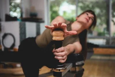 Leah is looking up and away from her hands which are placed on the GYROTONICs knob handle as she twists her spine to the left. A bright studio is in the background.