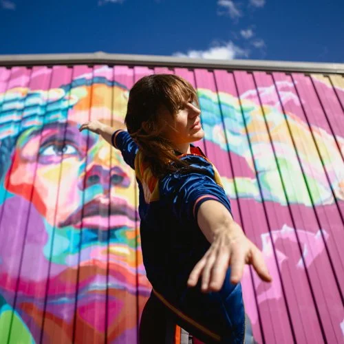 Leah stands in front of a building with a purple mural and blue sky on a sunny day. She is gazing into the distance and her arms are outstretched wide.