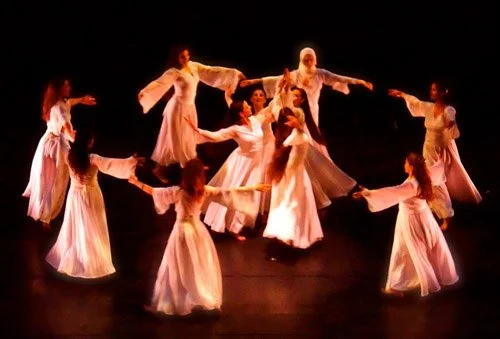Ensemble of dancers in flowing white dresses performing in a circle on a dark stage.