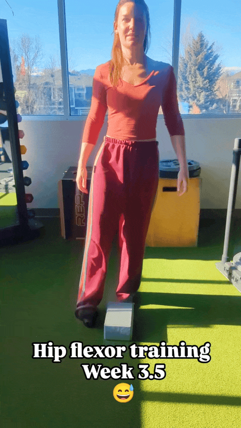 Leah is standing on one leg in a physical therapy studio with natural lighting, lifting her bent leg over a block with a small weight on her foot. Text overlay reads "Hip flexor training week 3.5" with a nervous sweating emoji.