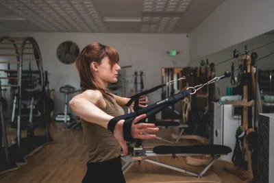Leah is pictured in a wide shot of a GYROTONICS studio in profile view, standing, with her elbows bent and hands in black straps of the pulley tower equipment.