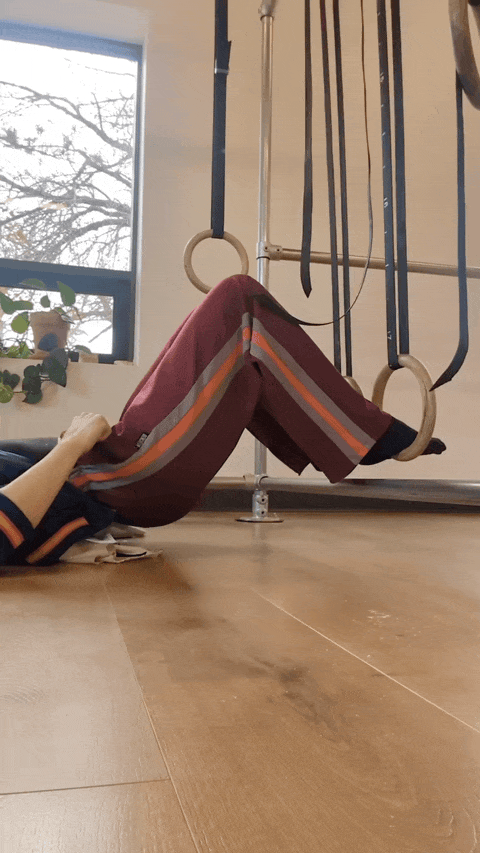Leah is laying on the floor with her feet in gymnastics rings, bending her knees, doing hamstring curls in a well-lit movement studio with wood floors.