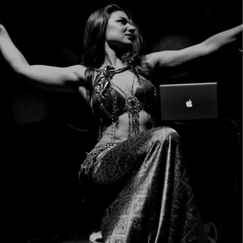 Leah a belly dancer in ornate costume with arms outstretched, seated next to a laptop on stage.