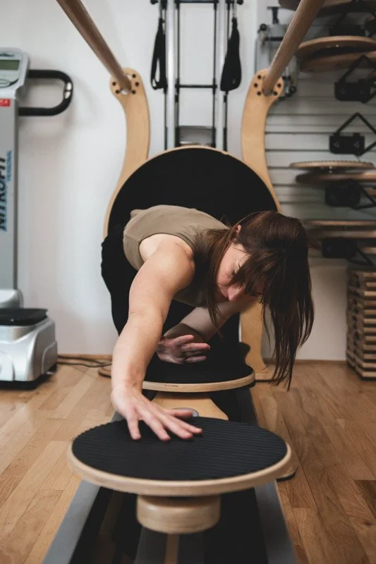 Leah is in a tank top and black pants doing GYROTONICs on a Jump Stretch Board with a wooden frame and black cushioning.