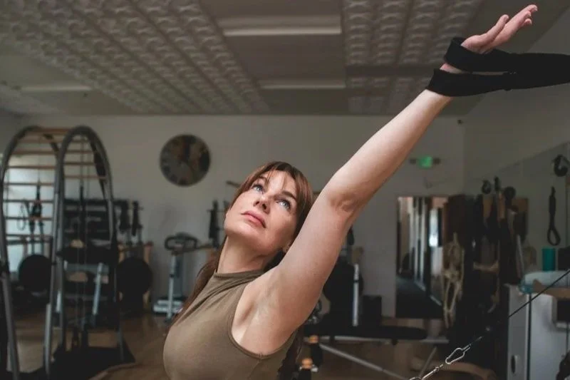Leah stretches her right arm overhead in a GYROTONICS® studio, with her wrist wrapped in a supportive strap.