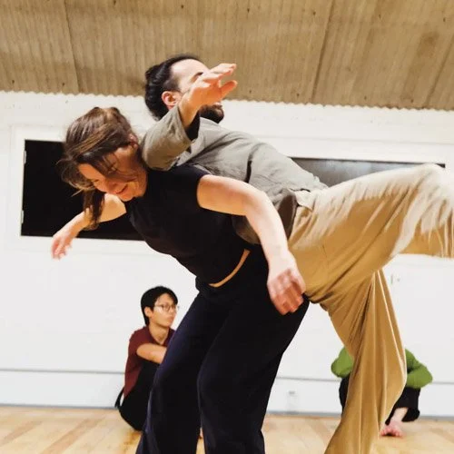 Leah is supporting her partner as they lean back-to-back in a contact improvisation class, with two other individuals observing in the background.