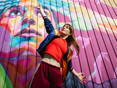 Leah a woman with her arms outstretched dances in front of a vibrantly colored mural depicting a large, abstract face.