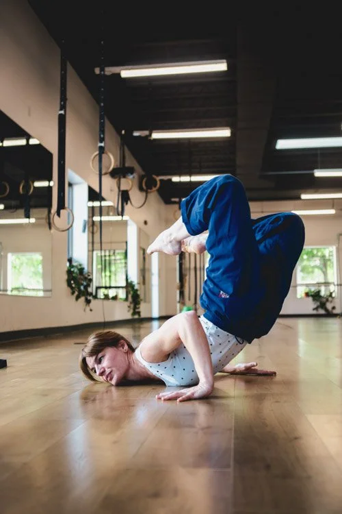 Leah is in a white tank top and dark blue pants performing an acrobatic roll and floorwork move on a wooden floor in a gym. Her chest and hands roll on the floor with legs overhead.