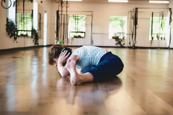Leah wears a white polka dot shirt and dark blue pants sitting on a wood floor in a gym. Her legs are extended in a middle split and upper body bent forward, holding a weight over her head.