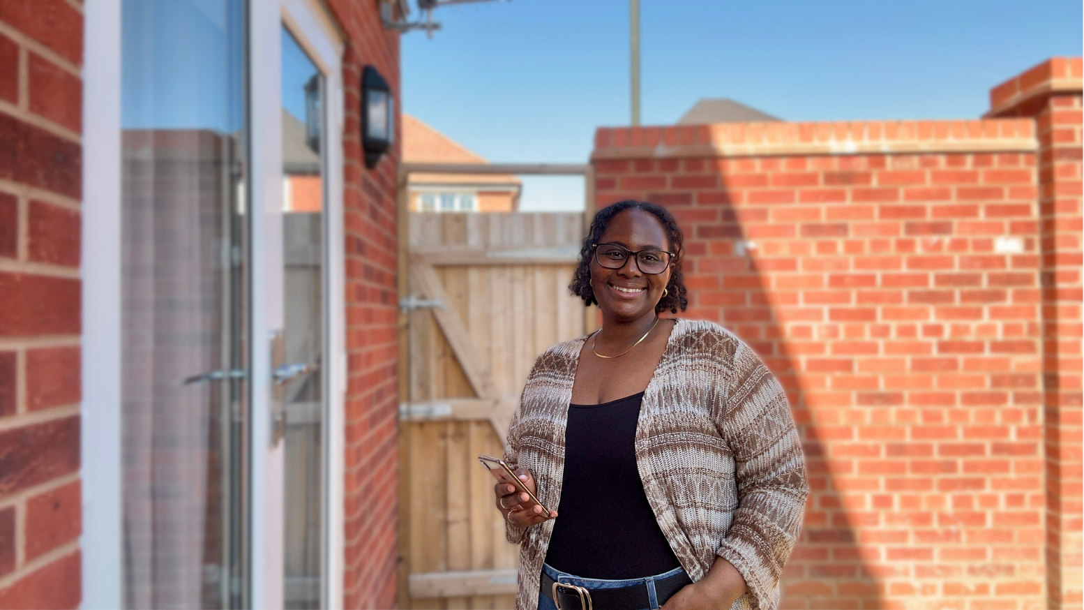 Destiny May the Business Owner standing outside in a backyard, smiling, holding a smartphone, with a brick wall and wooden gate in the background.