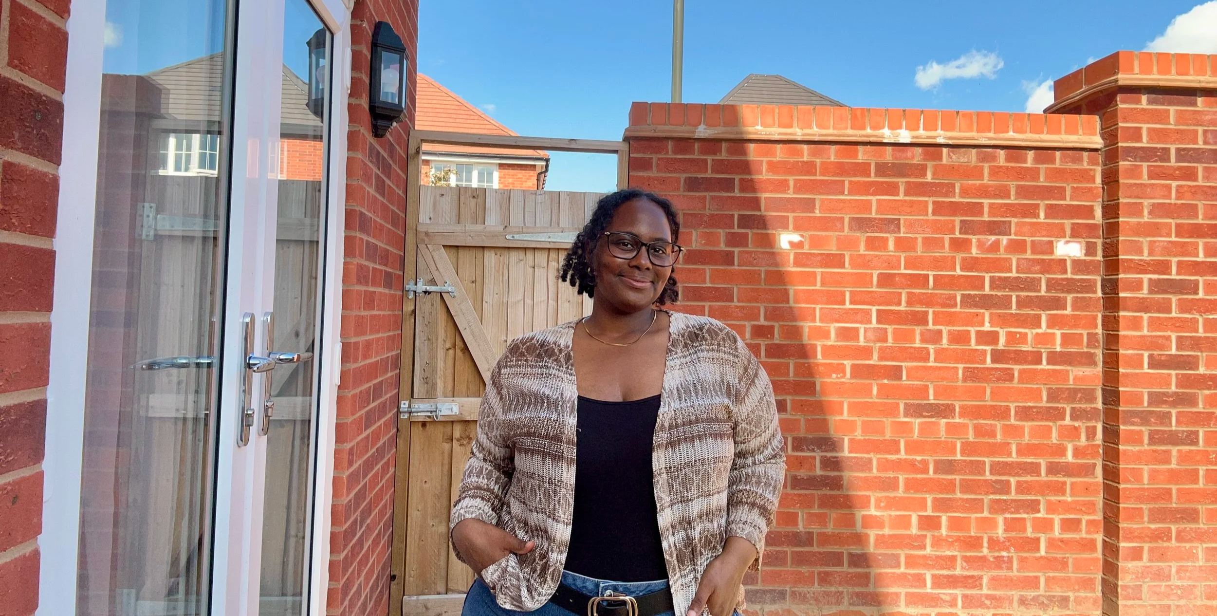 Destiny May the Business Owner with glasses and curly hair, wearing a patterned blazer and black top, standing outdoors in front of a brick wall and wooden gate, with a clear blue sky above.