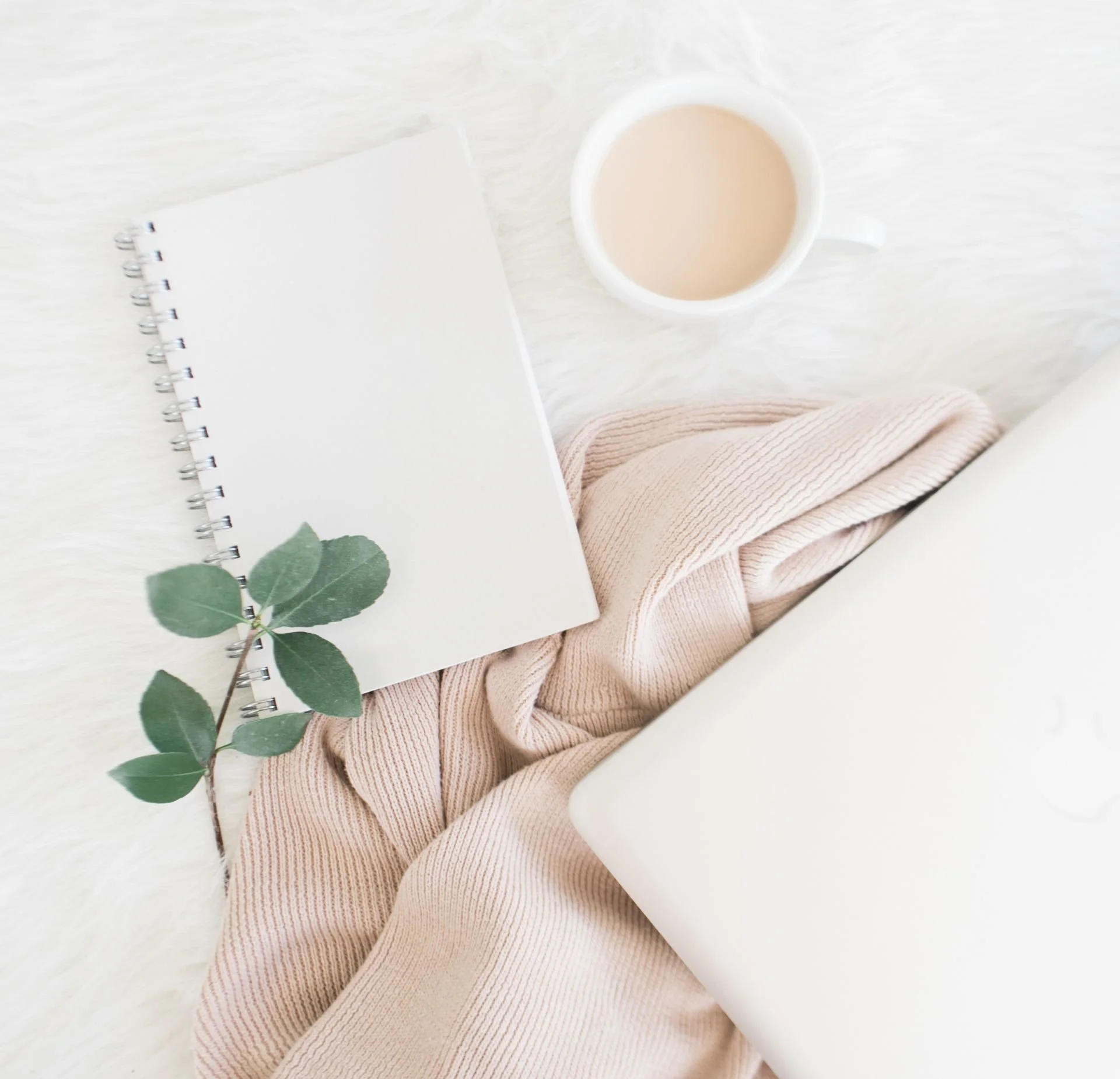 Overhead view of a white table with a spiral notebook, a cup of coffee with milk, and a beige sweater.