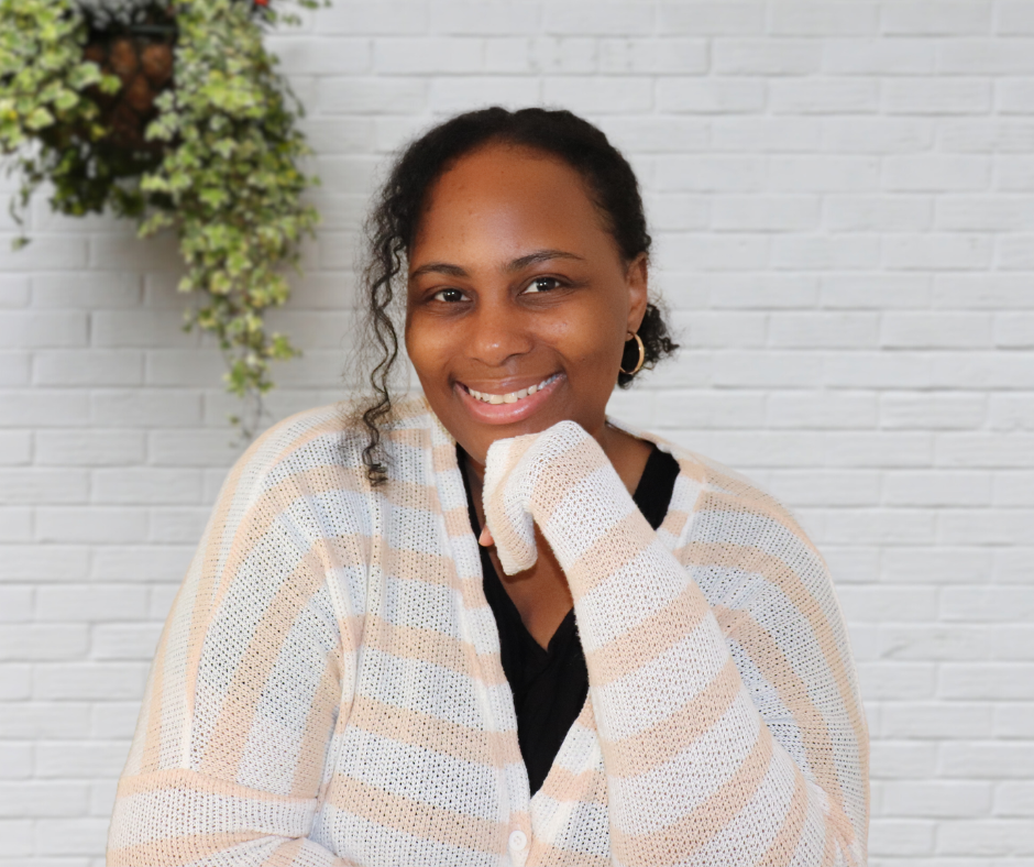 Destiny May the Business Owner is smiling with one hand resting under her chin, standing in front of a white brick wall with a green plant in the background.