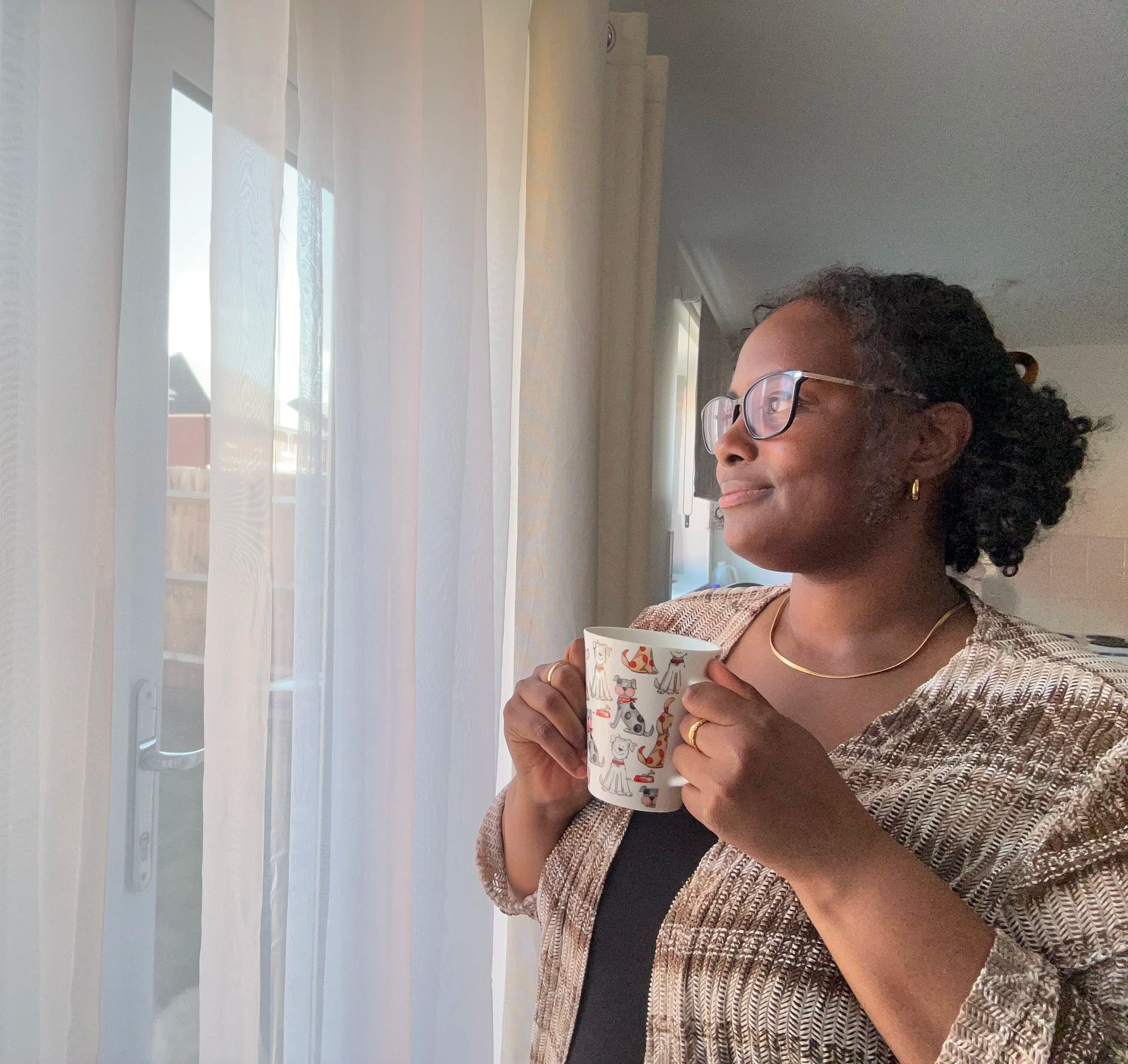 Destiny May the Business Owner with curly hair and glasses holds a mug with animal illustrations, sitting indoors near a window with sheer curtains, wearing a patterned blazer.