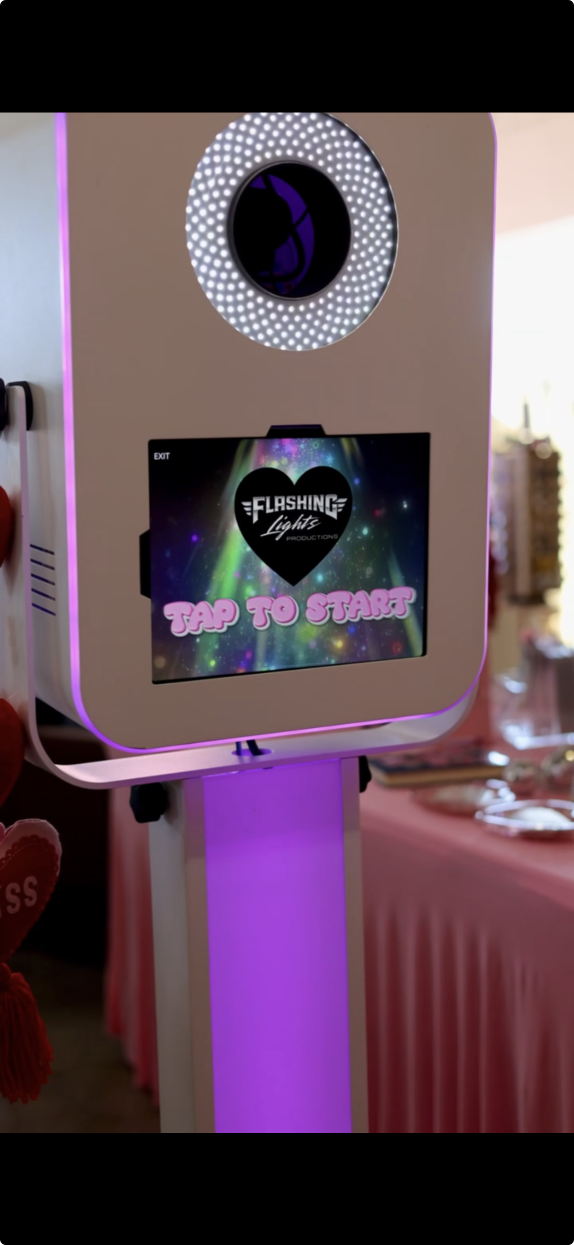 Photo of a photo booth with a pink frame and a screen displaying 'Flashing Lights Productions' and 'Tap to Start' with colorful lights and a heart logo.