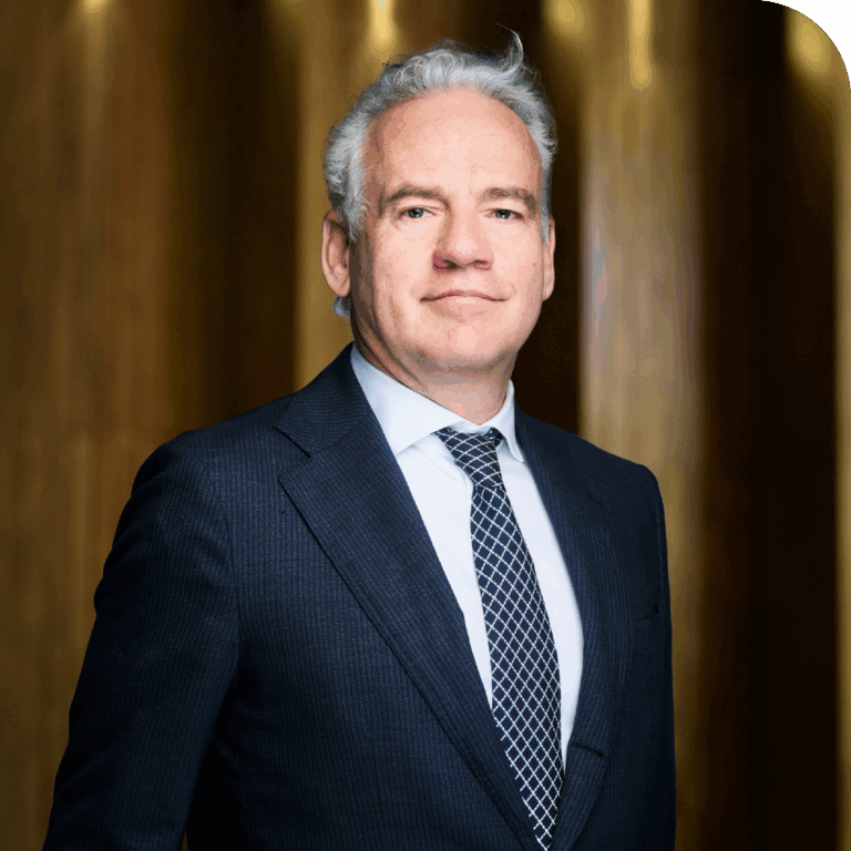 A middle-aged man with gray hair dressed in a dark suit, white shirt, and patterned tie, standing in front of a blurred wooden background.