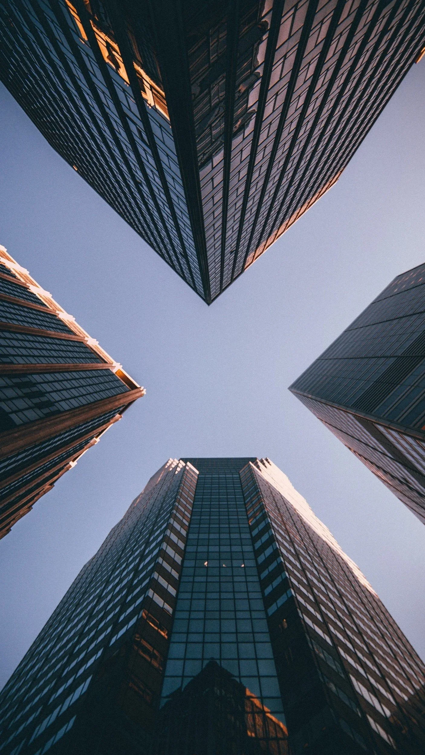 Looking up at tall glass skyscrapers in a city at dusk, reflecting the sunset light, with a clear sky above.