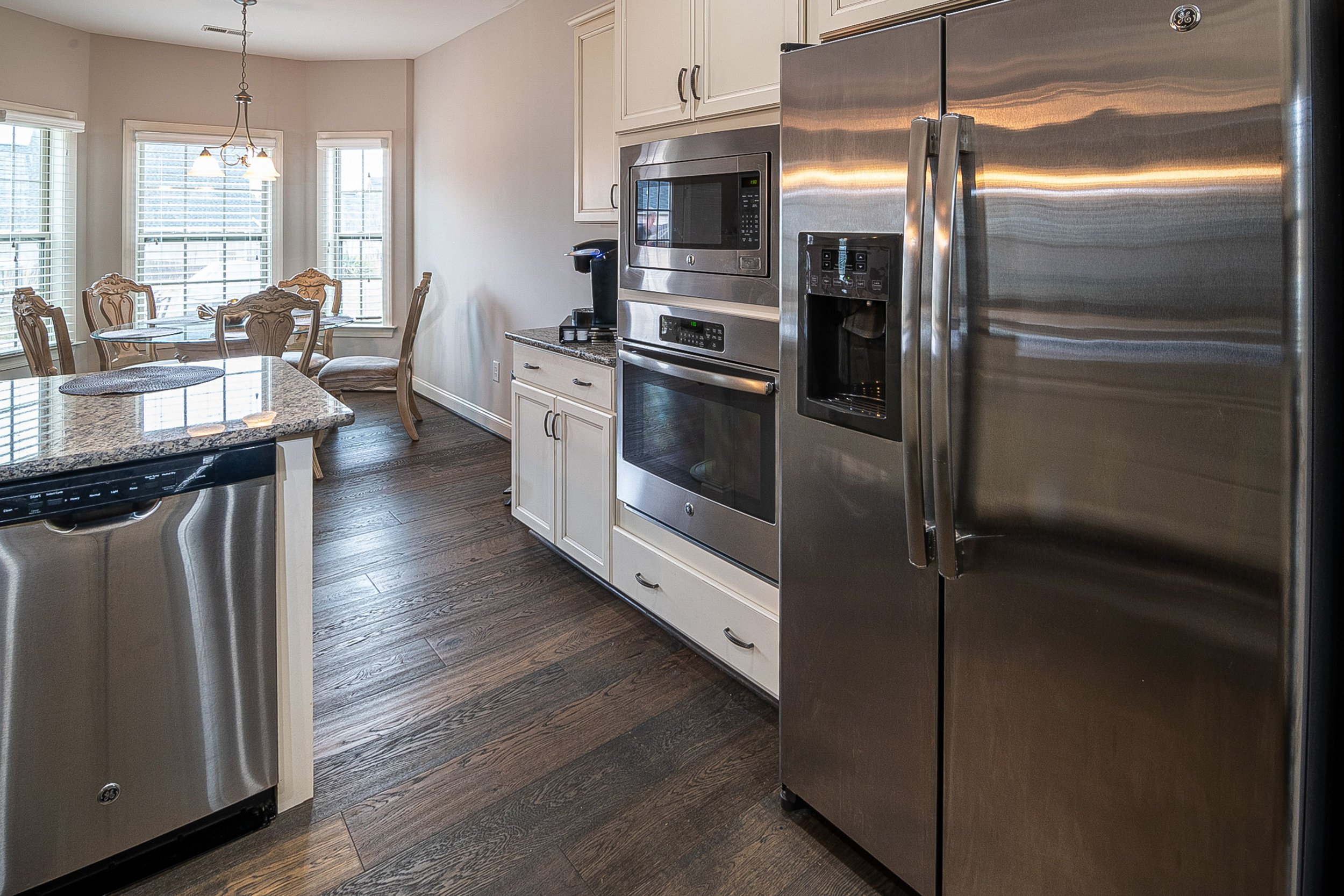Modern kitchen with stainless steel refrigerator, built-in microwave, white cabinets, and a dining area with a round glass table and ornate chairs near multiple windows. Dark wooden floor and a hanging light fixture.