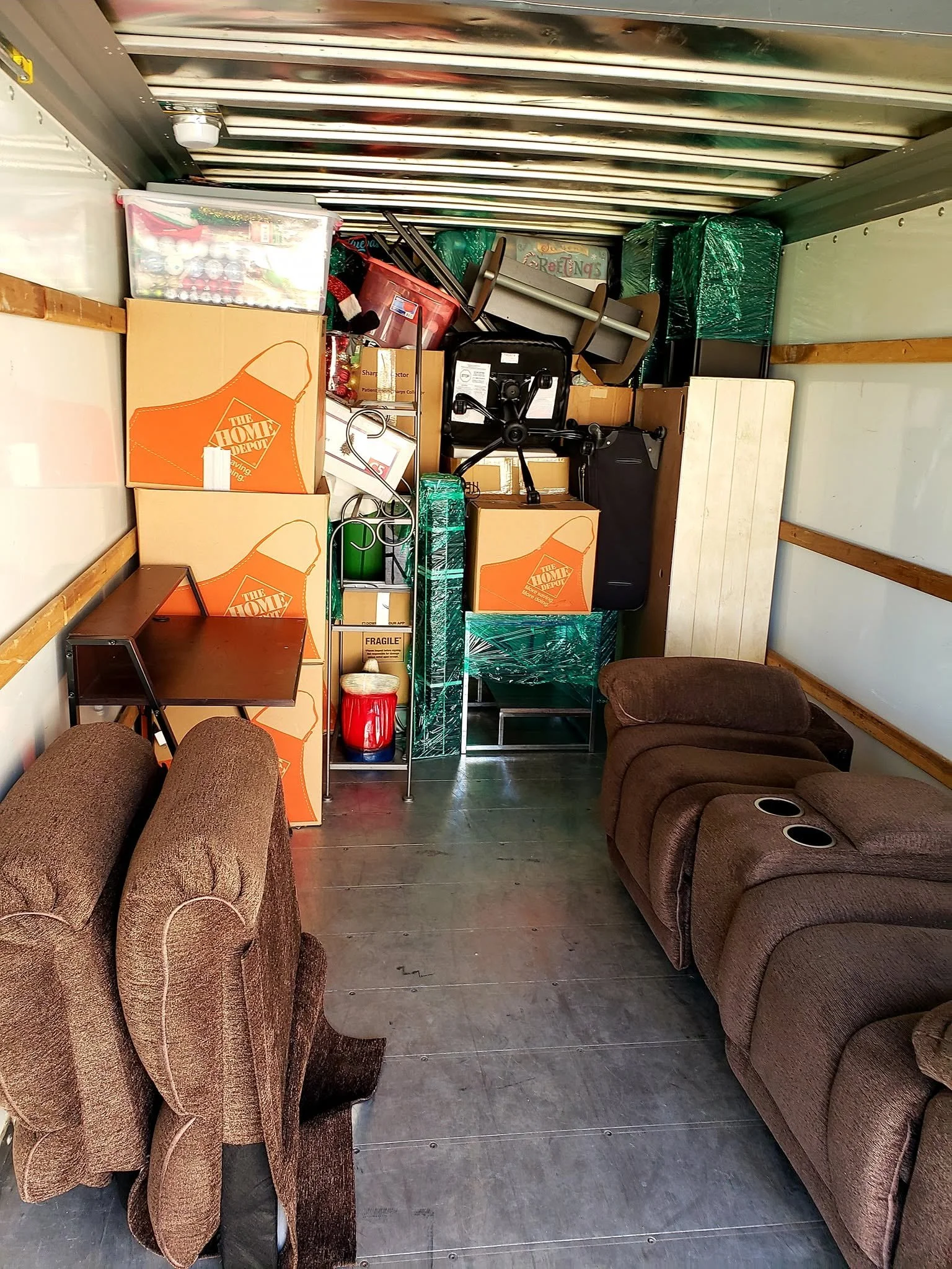 The interior of a moving truck filled with furniture, boxes, and storage containers. There are brown sofas, cardboard boxes, plastic storage bins, and some office chairs stacked against the back wall. The floor is made of wood planks and some objects
