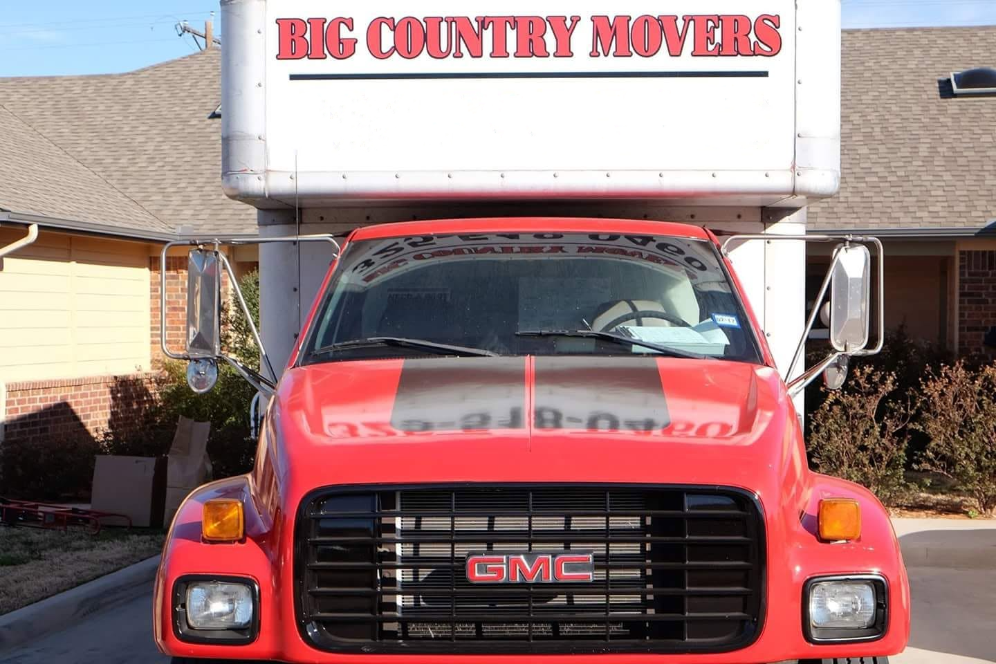 Red GMC moving truck with large box on top, parked in front of a residential house with beige siding and brick foundation.