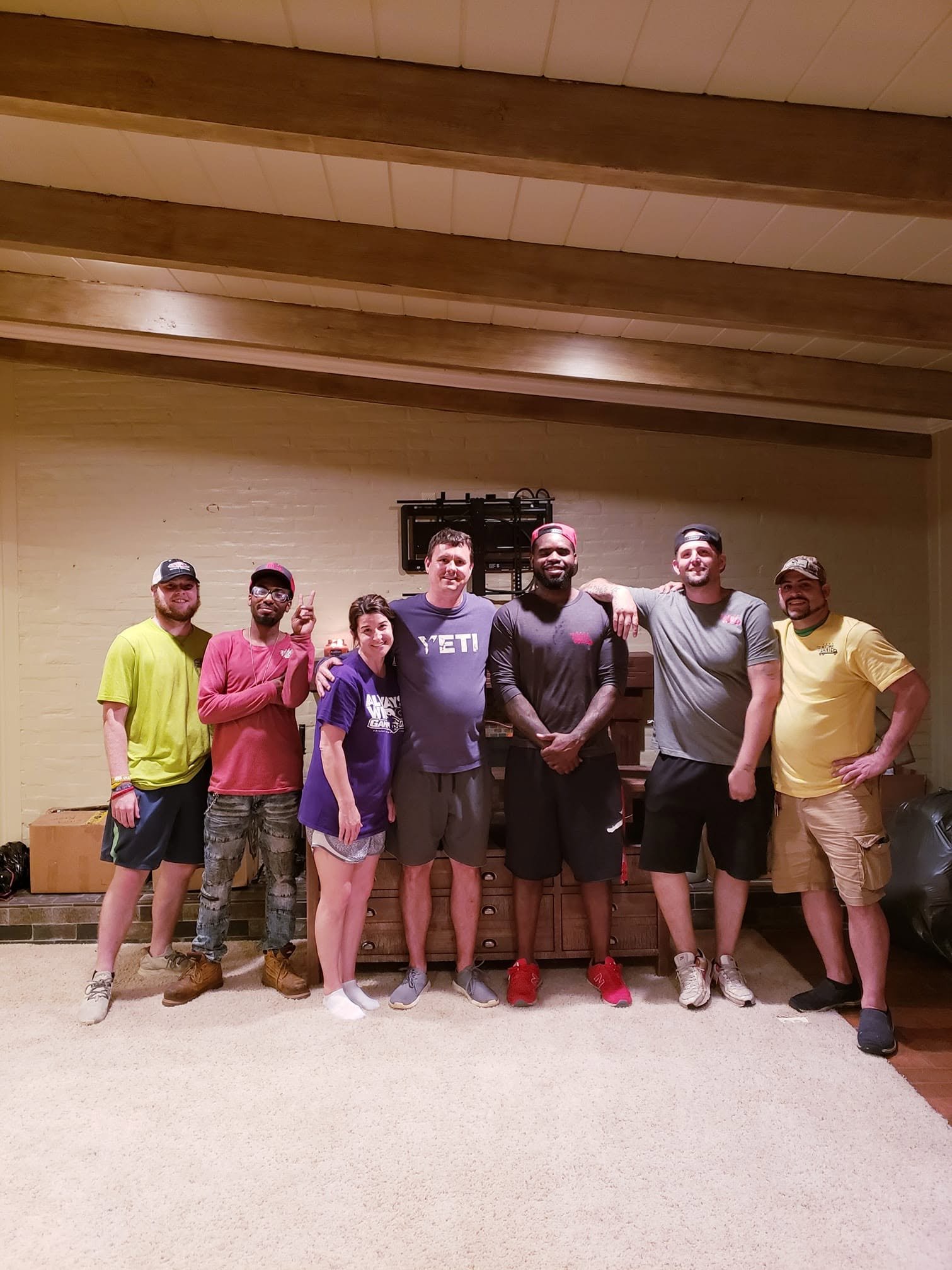 Group of eight people standing together indoors, smiling for the camera. They are casually dressed and appear to be friends or family, standing in front of a wooden cabinet and a beige brick wall.