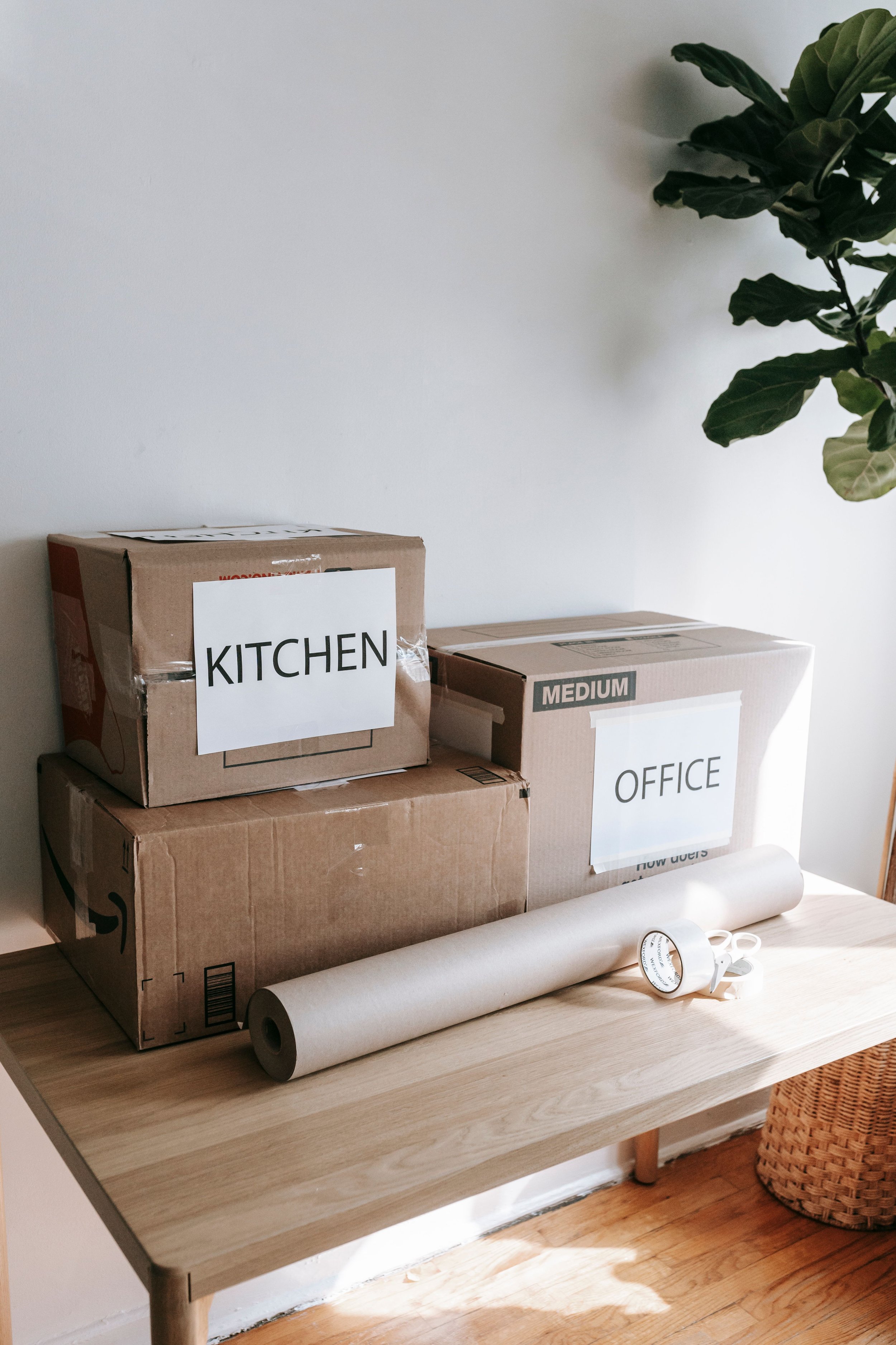 Boxes labeled 'KITCHEN' and 'OFFICE' stacked on a light wooden table, with a roll of white wrapping paper, scissors, and a white wristwatch nearby, near a wicker basket and a green leafy plant.