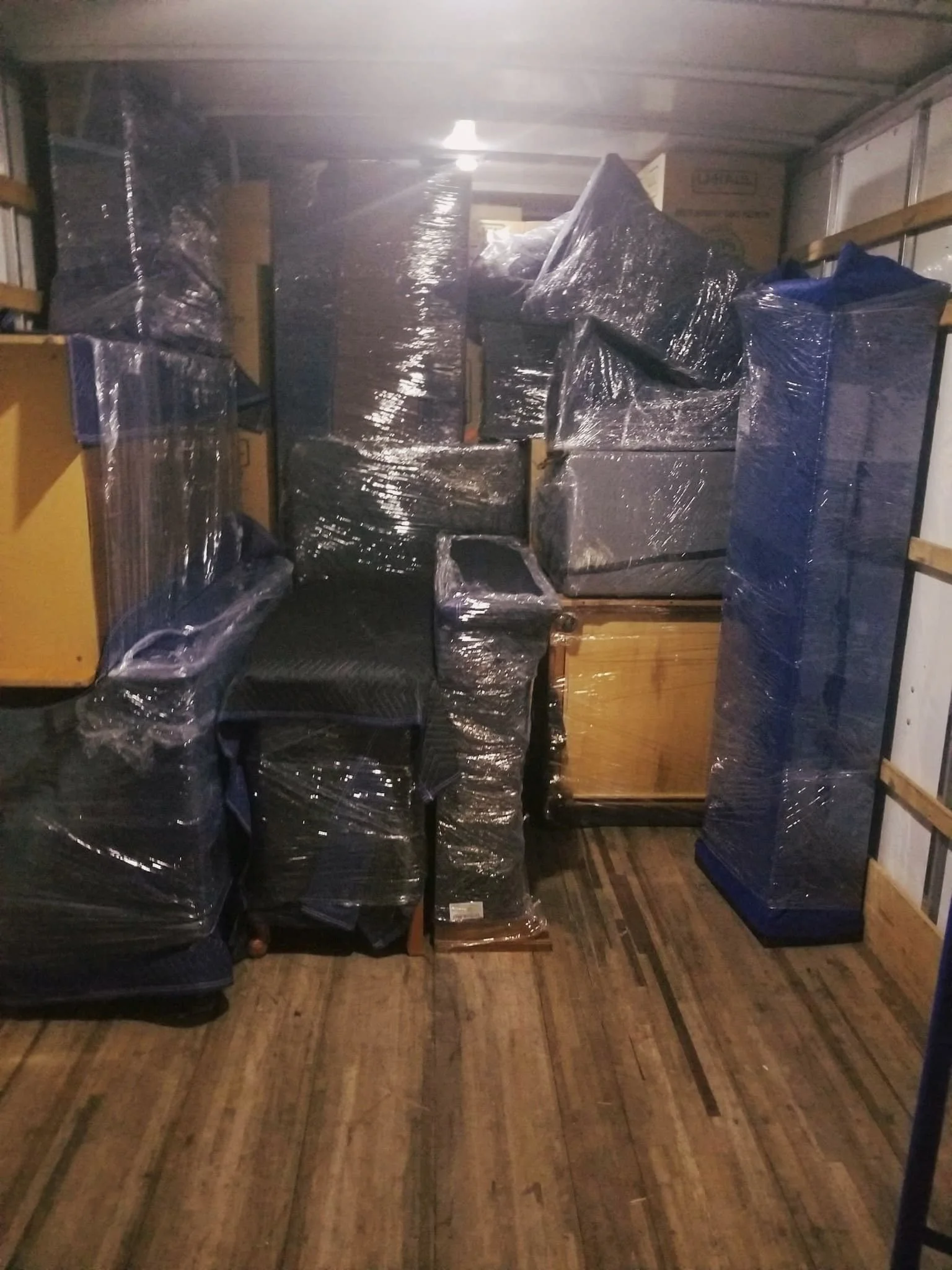 A storage area or warehouse with cardboard boxes, furniture wrapped in plastic, and wooden shelving units, all stored inside a metal container or truck with lower wooden flooring.