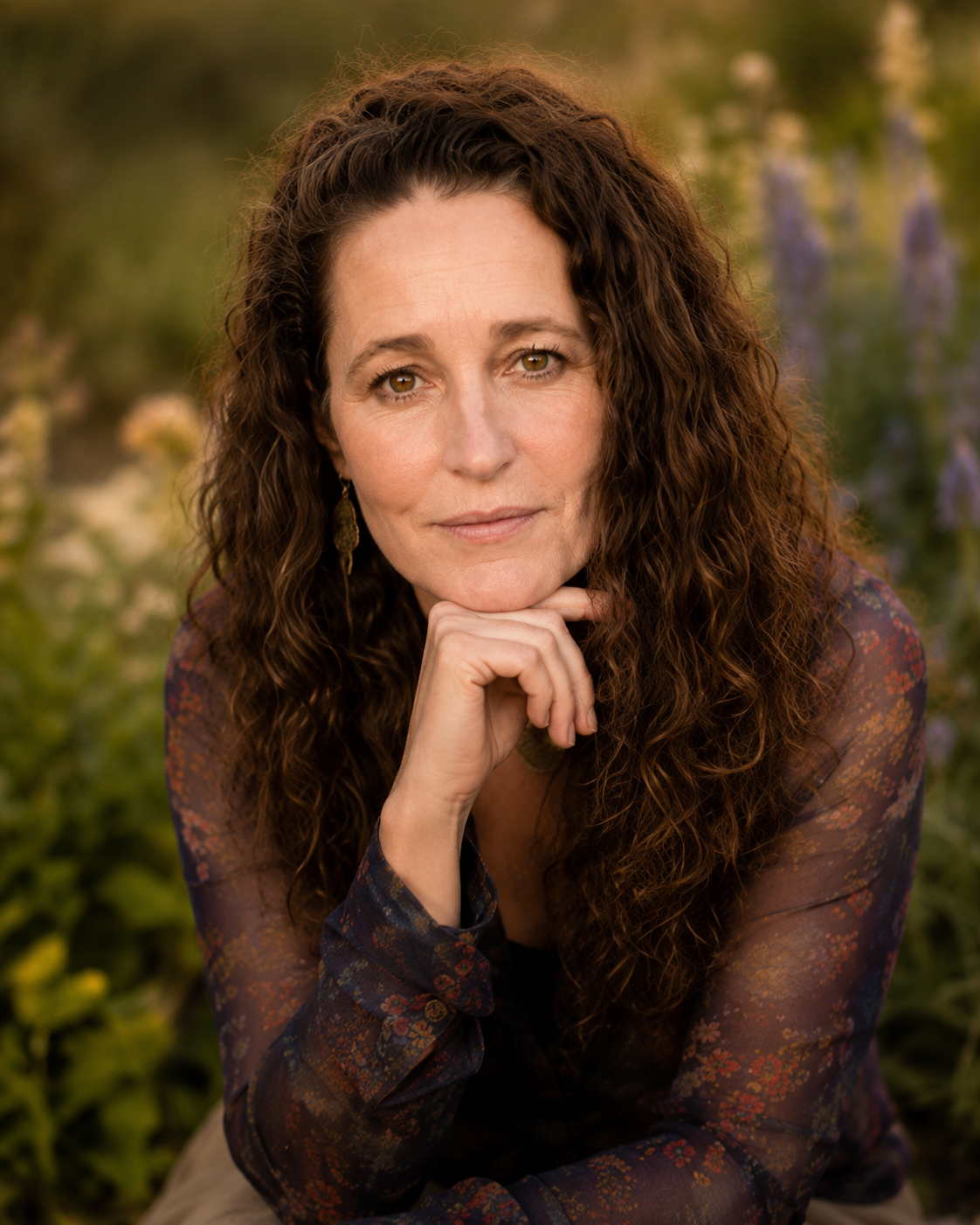 A woman with curly brown hair and brown eyes resting her chin on her hand outdoors, surrounded by blurred green and purple foliage.